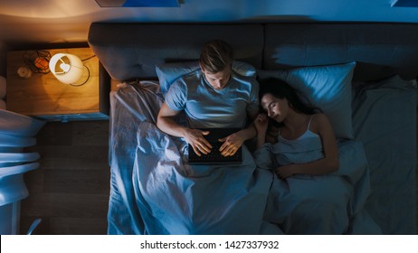 Top View Young Man in Bed Working on a Laptop Computer at Night while Girflfriend Sleeps Beside Him. Exceptionally Dedicated Project Manager Finishing Work in Bed at Night. - Powered by Shutterstock