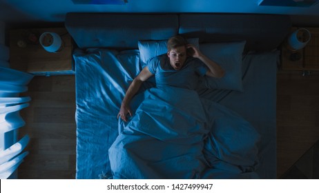 Top View Of A Young Man In Bed At Night Having Terrible Nightmare, He Wakes Up Scared And Covered In Sweat.