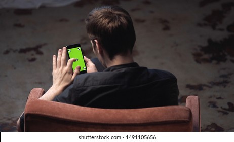 Top View Of Young Man With Brown Hair Sitting In Chair And Tapping On IPhone Green Screen In Dark Room. Stock Footage. Chroma Key