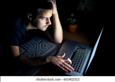 Top view of young man reading news on laptop lying in dark room. Looking concentrated