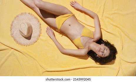 Top view of young pretty fit brown-haired Asian woman in yellow crochet swimwear lies down on yellow terry blanket and takes a sunbath | UV protection and body care commercial