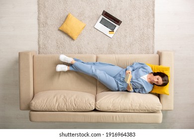 Top view of young restful brunette female in blue pajamas reading book while lying on couch with head on yellow pillow after working day - Shutterstock ID 1916964383