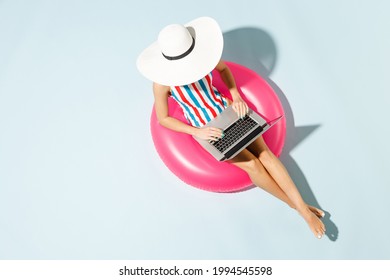 Top view young sexy woman slim body wear striped one-piece swimsuit hat sit inflatable tube ring hotel pool work on laptop pc isolated on pastel blue color background. Summer sea rest sun tan concept