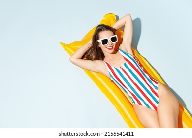 Top view young sexy woman slim body wear striped one-piece swimsuit sunglasses lies on inflatable mattress hotel pool isolated on pastel blue color background. Summer vacation sea rest sun tan concept