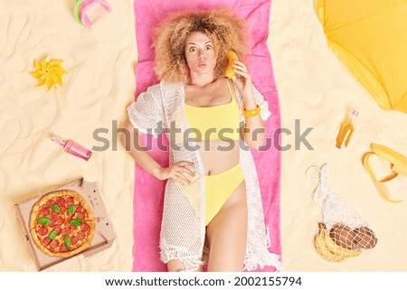 Top view of young slim woman with curly bushy hair keeps banana near ear as of telephone dressed in bathing suit lies on towel spends summer holidays at seaside surrounded by beach acessories