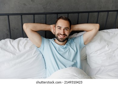 Top View Young Smiling Man Wear Casual Blue T-shirt Lying In Bed Hands Folded Under Head Look Camera Rest Relax Spend Time In Bedroom Lounge Home In Own Room House Wake Up Dream. Real Estate Concept