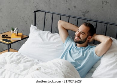 Top View Young Smiling Man Wear Casual Blue T-shirt Lying In Bed Hands Folded Under Head Look Camera Rest Relax Spend Time In Bedroom Lounge Home In Own Room House Wake Up Dream. Real Estate Concept