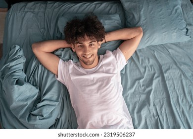 Top view young smiling man wear white t-shirt lying in bed hands folded under head look at camera, spend relax time in bedroom, wake up dream. Copy space - Powered by Shutterstock