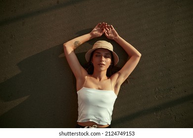 Top View Of Young Stylish Female In Bucket Hat And Tank Top Lying With Closed Eyes On Asphalt Ground On Sunny Day In Summer