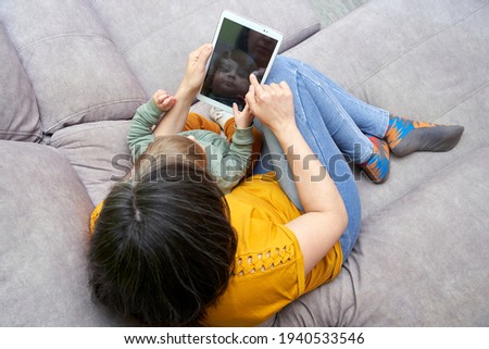 Top view of young woman with her daughter sitting on the sofa at home using a tablet. Concept of family time, gadgets, conciliation, lifestyle.