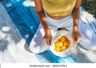 Top view of a young woman holds white headwear with lemons. Girl in white pants and yellow jersey holding a cotton basket of fresh-picked lemons sitting on the picnic blanket on a sunny day outdoors. - Powered by Shutterstock