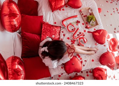 Top view of young woman sitting on bed around red balloons, rose petals. Faceless blogger holds heart-shaped beads. Decoration with inscription Love, pillows, wooden tray with two glasses and grapes. - Powered by Shutterstock