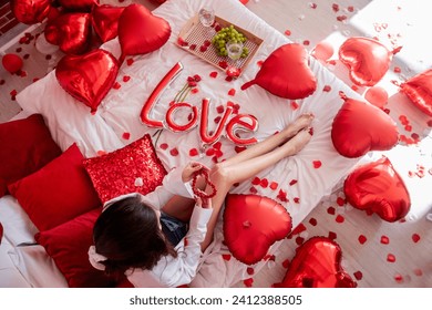 Top view of young woman sitting on bed around red balloons, rose petals. Faceless blogger holds heart-shaped beads. Decoration with inscription Love, pillows, wooden tray with two glasses and grapes. - Powered by Shutterstock