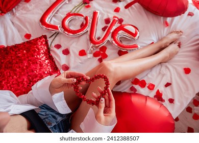 Top view of young woman sitting on bed around red balloons, rose petals. Faceless blogger holds heart-shaped beads. Decoration with inscription Love, pillows, wooden tray with two glasses and grapes. - Powered by Shutterstock