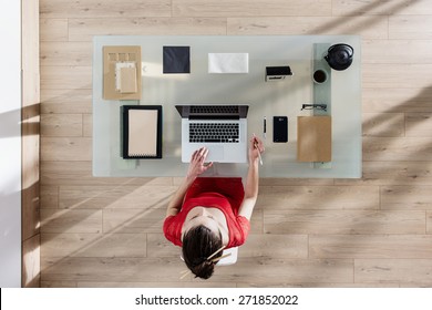 Top View, Young Woman Sitting At Glass Desk And Working On Her Laptop, Her Table Is Perfectly Tidy, The Sun Casts Graphics Shadows On The Wood Floor