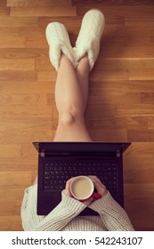 Top View Of A Young Woman Sitting On The Living Room Floor, Leaning Against A Couch, Drinking Coffee And Using A Laptop Computer