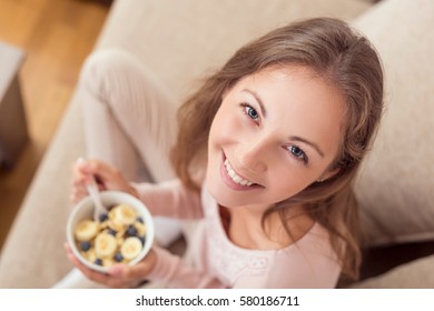 Top View Of A Young Woman Sitting On A Living Room Couch, Holding A Bowl Of Cereal And Having Breakfast