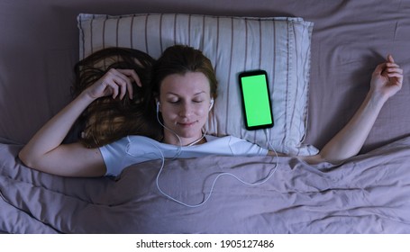Top view of young woman sleeping with earphones and smartphone with green screen. Audio books apps, sleep control app.
