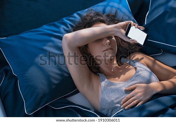 Top view of young woman sleeping in bed while\
holding mobile phone. Girl sleeping in blue linen with smart phone.\
Woman lying in bed fell asleep while texting meassage with\
smartphone late in night.