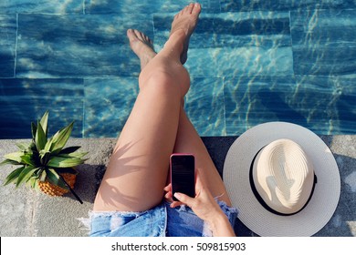 Top View Young Woman Sunbathing Near Swimming Pool Edge Using Her Smart Phone. Relaxing On Summer Holiday With Pineapple Cocktail