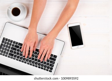 Top View Of Young Woman Surfing Internet, Online Shopping, Close Up Of Hands On Her Laptop Keyboard On White Wood Textured Office Desk. Overhead Shot Female At Workspace, Coffee, Cell Phone, Supplies.