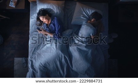 Top View: Young Woman Uses Smartphone in Bed at Night When Her Male Partner Trying to Fall Asleep Beside. Couple Fight, Argue. Addictive World of Social Media, Doom Scrolling, Fake News.