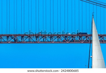 Top of the white sail of a sailboat sailing in front of the red steel 25 de Abril suspension bridge with road traffic and a train traveling inside the bridge under a clear blue sky.