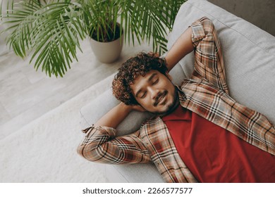 Top with young Indian man wearing casual clothes close eyes take nap sleep lay down on grey sofa couch stay at home hotel flat rest relax spend free spare time in living room indoor. Lounge concept