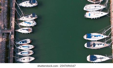 Top-down aerial view of a marina with neatly docked sailboats and yachts lined along calm green waters. The organized layout of the vessels and the pier creates a striking geometric composition. - Powered by Shutterstock