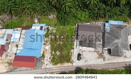 Top-down aerial view of an undeveloped vacant land lot surrounded by residential houses, a local road and tropical palm plantation in Malaysia.