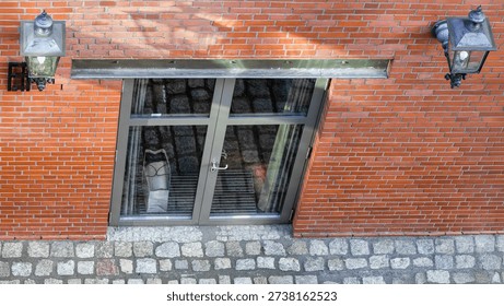 Top-down angled view of a red brick building facade with basement glass door and paper shopping bag visible inside. Urban architectural detail with vintage wall lanterns and cobblestone pavement in natural daylight.