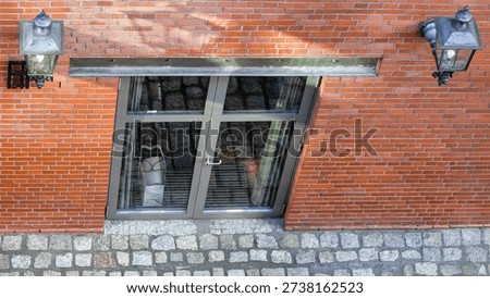 Top-down angled view of a red brick building facade with basement glass door and paper shopping bag visible inside. Urban architectural detail with vintage wall lanterns and cobblestone pavement in natural daylight.