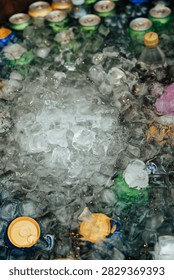 Top-down close-up of assorted beverage cans surrounded by melting ice cubes in a cooler, emphasizing cold refreshment at an outdoor event