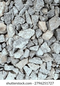 Top-down close-up shot of angular crushed stone aggregate, featuring mixed shades of gray, off-white, and light brown. Natural rough texture of granite gravel, commonly used in construction, road base, concrete mixing, landscaping, and outdoor paving. Detailed surface pattern with varying stone sizes, suitable as a background or material reference.