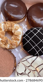A top-down, close-up shot of a half-dozen assorted gourmet donuts neatly arranged inside a white delivery box. The variety includes classic chocolate glazed donuts, a white frosted ring donut generously topped with sliced toasted almonds, a dark chocolate cookie crumb donut with a white icing grid pattern, a cocoa powder-dusted chocolate ring donut, and a white chocolate marbled donut with dark swirls. Bright, natural lighting highlights the glossy textures of the glazes and toppings.