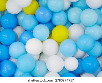 ​A top-down, close-up view of a ball pit filled with numerous colorful plastic spheres. The palette is dominated by various shades of blue and clean white, with occasional bright yellow balls providing a cheerful and energetic contrast.