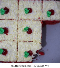 Top-down, close-up view of a sliced red velvet sponge cake topped with a layer of buttercream and a generous coating of grated cheddar cheese. Each square slice is neatly decorated with a small red glazed cherry topping and a green buttercream leaf accent. One slice has been removed from the grid, revealing a corner of the plain cardboard box base. The shot is captured with a soft focus, highlighting the rich textures of the shredded cheese and the vibrant contrast of colors, making it ideal for culinary, bakery, or dessert-themed content.