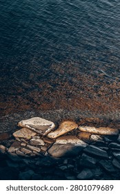 Top-down close-up view of smooth weathered stones at the water’s edge. Transparent dark blue sea, underwater vegetation, warm sunlight on wet rocks, natural coastal texture and calm atmosphere.