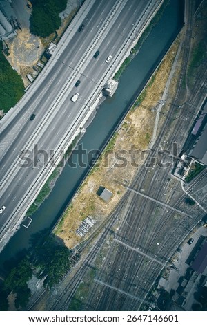 Top-down drone view of multilane highway with moving vehicles, railway yard with numerous tracks, and a narrow canal running between them. Complexity of modern transportation networks