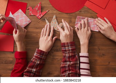 Top-down flat lay shot of unrecognizable woman and two girls sitting at wooden table using colored paper to make origami cranes