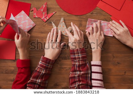 Top-down flat lay shot of unrecognizable woman and two girls sitting at wooden table using colored paper to make origami cranes