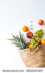 A top-down flat lay of a straw basket overflowing with fresh fruit, including pineapple, green grapes, peaches, and oranges, resting on a white surface.