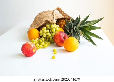A top-down flat lay of a straw basket overflowing with fresh fruit, including pineapple, green grapes, peaches, and oranges, resting on a white surface.