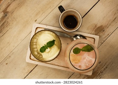 Top-down flat lay view of a cafe beverage and dessert pairing featuring scoops of creamy vanilla ice cream topped with a fresh green mint sprig in a fluted glass, a cup of dark espresso coffee, and a cold iced pink fruit drink garnished with mint and lychee, arranged on a white wooden tray over a rustic tabletop surface.