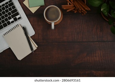 Top-down flat lay view of a warm, rustic wooden desktop workspace featuring a silver laptop, a small spiral notepad with a black pen, and a white coffee mug on a coaster. The background is accenting by sticky notes, a holder filled with wooden pencils, and green plant leaves in the corner, leaving plenty of empty copy space on the dark wood surface—ideal for creative writing blogs, professional branding, or cozy remote work themes.