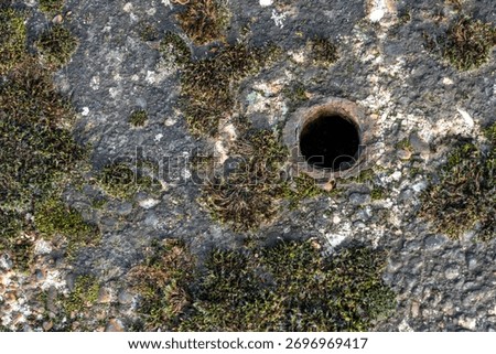 Top-down, full frame close-up of a circular rusted metal pipe hole situated slightly off-center on a heavily textured concrete surface covered by dark moss and pebbles