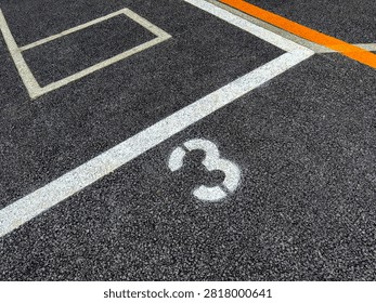 A top-down, high-angle close-up shot capturing a white stenciled number 3 painted on fresh, dark textured asphalt pavement inside a designated parking space. White and orange painted boundary lines crisscross the road surface, creating geometric lines and angles. Ideal for stock media on transportation, parking lot management, road markings, urban infrastructure, numbers, logistics, and asphalt textures.