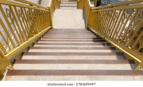 A top-down, high-angle shot looking down a pedestrian staircase on the Sixth Street Bridge (Roberto Clemente Bridge) in Pittsburgh, Pennsylvania. The image highlights the concrete steps detailed with textured, slightly rusted metal safety treads, flanked by the bridge's signature Aztec Gold painted steel railings with ornate, arched geometric designs. Bright midday sunlight casts crisp shadows across the walkway, capturing the industrial charm and architectural details of this iconic Three Sisters bridge.