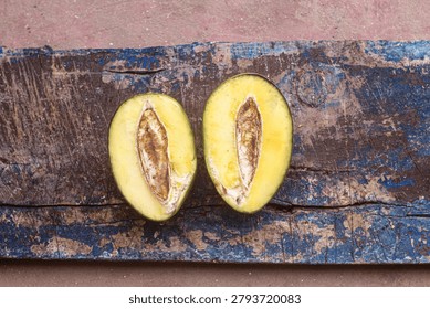 Top-down, horizontal view of a single raw green mango cut into halves, revealing the crisp yellowish inner flesh and central seed pit. The tropical fruit slices rest on a rustic, heavily weathered wooden plank with peeling blue paint, creating a striking contrast for themes of rural agriculture, food preparation, and textured design backdrops.