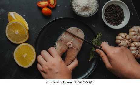 Top-down image of hands slicing fresh mackerel on a black plate, surrounded by ingredients like lemon slices, cherry tomatoes, rosemary, garlic, sea salt, and black pepper. - Powered by Shutterstock